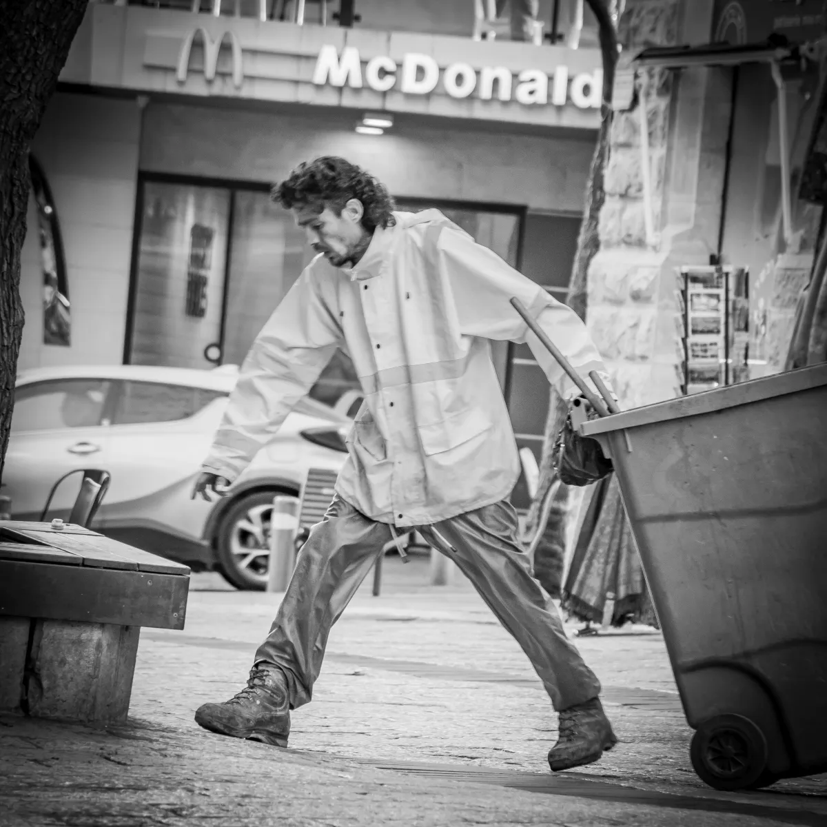 Man in a loose raincoat striding past a trash bin beneath a McDonalds sign